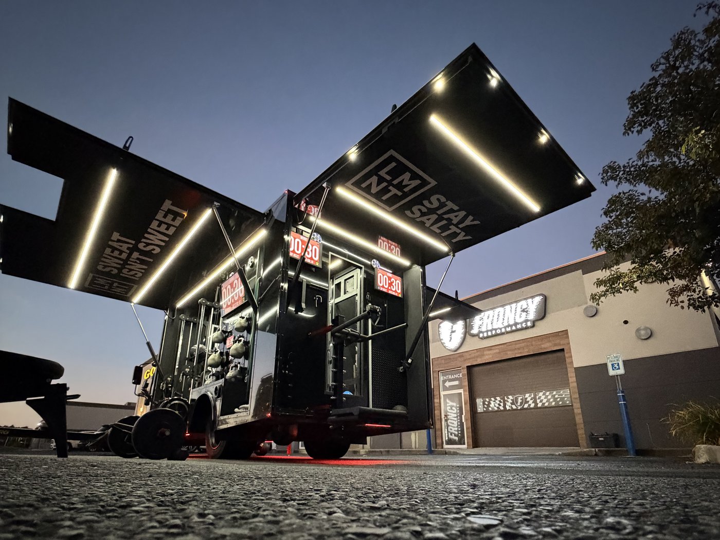 LMNT-branded Fit Truk mobile gym trailer at dusk with its gull-wing awnings raised, LED-lit training equipment and red interval timers inside