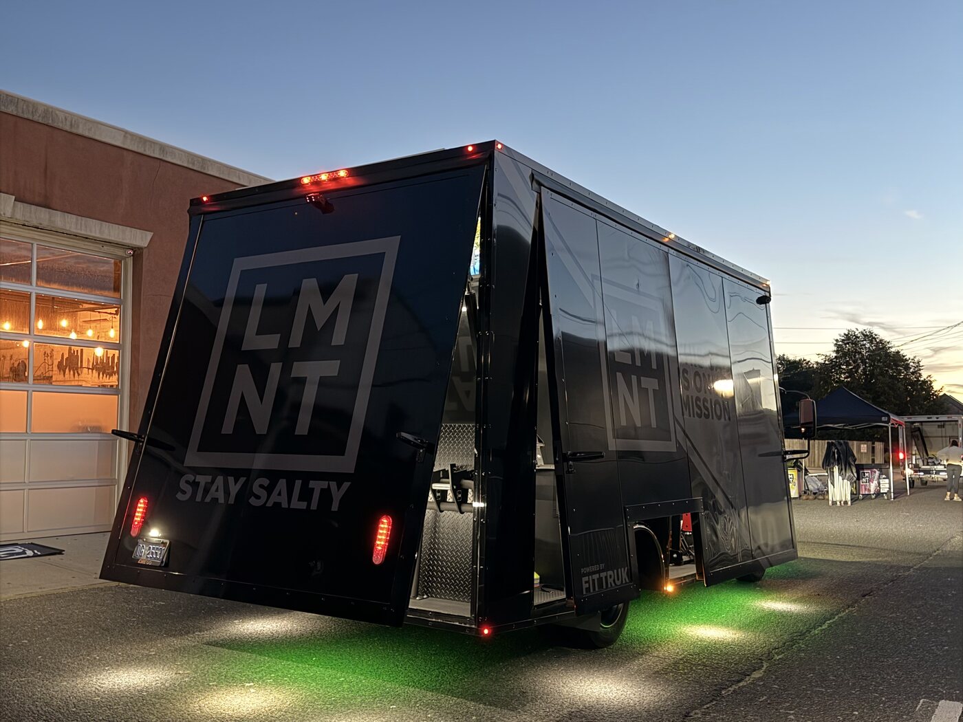 LMNT-branded Fit Truk mobile gym trailer at dusk on an asphalt lot, its side raised to show the training interior, green LED underglow pooling beneath it and warm puddle lights on the ground