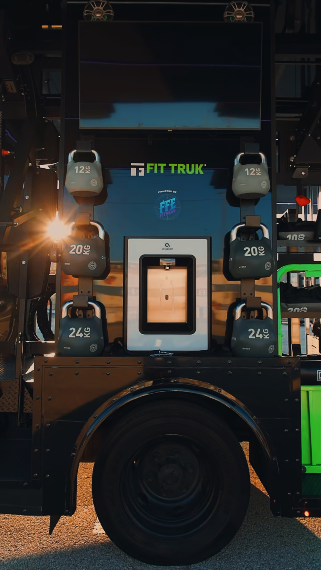 Close view above the rear wheel well of kettlebells in 12, 20 and 24 kg flanking a recessed Avalon bottle-refill station, with a blank display screen and two overhead speakers