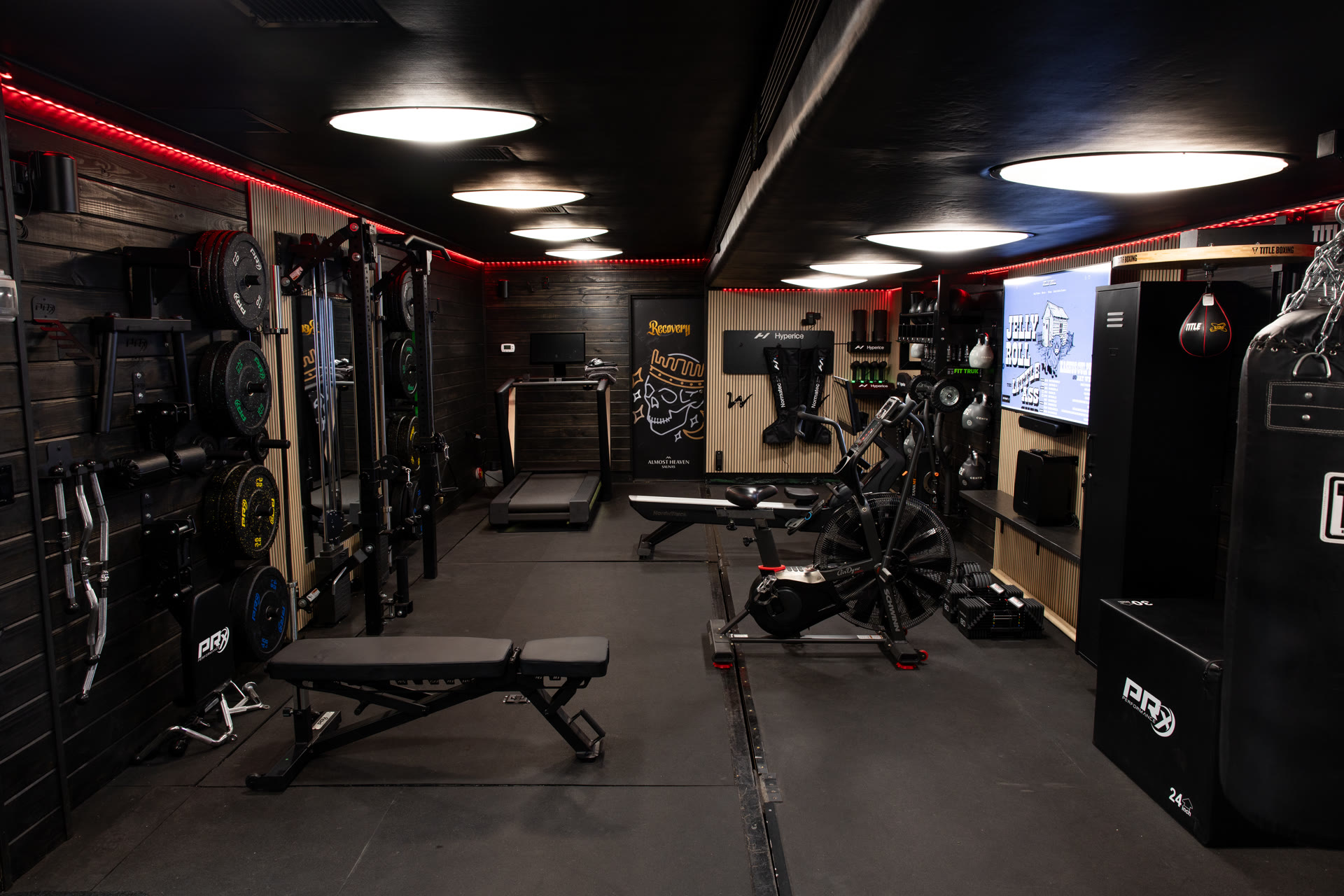 Custom gym trailer interior looking down the training corridor, weight racks and barbells on the left, treadmill at the rear, boxing bags and recovery gear on the right, red LED accent lighting