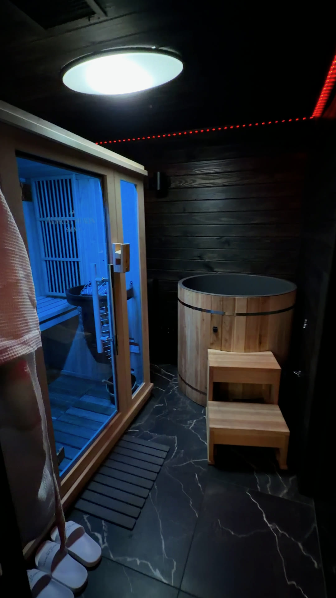 Custom mobile wellness build: wood sauna cabin glowing blue beside a wood cold plunge tub, white robe and slippers on a dark marble floor
