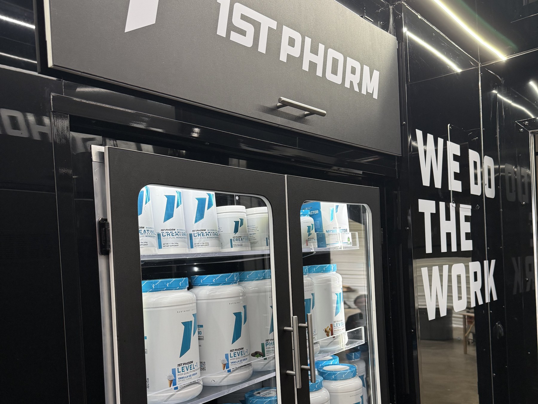 Interior of a custom Fit Truk build: a glass-door display refrigerator stocked with supplement tubs, set beside a wall reading 'WE DO THE WORK'