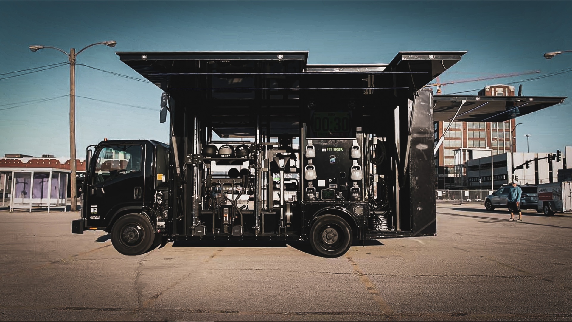 Opposite side of the deployed FFE Fit Truk 2.0 with a power rack and pull-up frame, medicine balls on shelves, kettlebells on wall pegs, dumbbells and plates on lower racks, and battle ropes stowed near the rear
